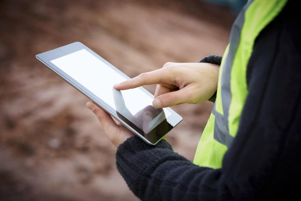 A person in a high-visibility vest uses a tablet outdoors, integrating holistic workforce management strategies. The background is blurred, suggesting a construction site. The individual interacts with the device, possibly streamlining tasks and coordinating efficient workflows.