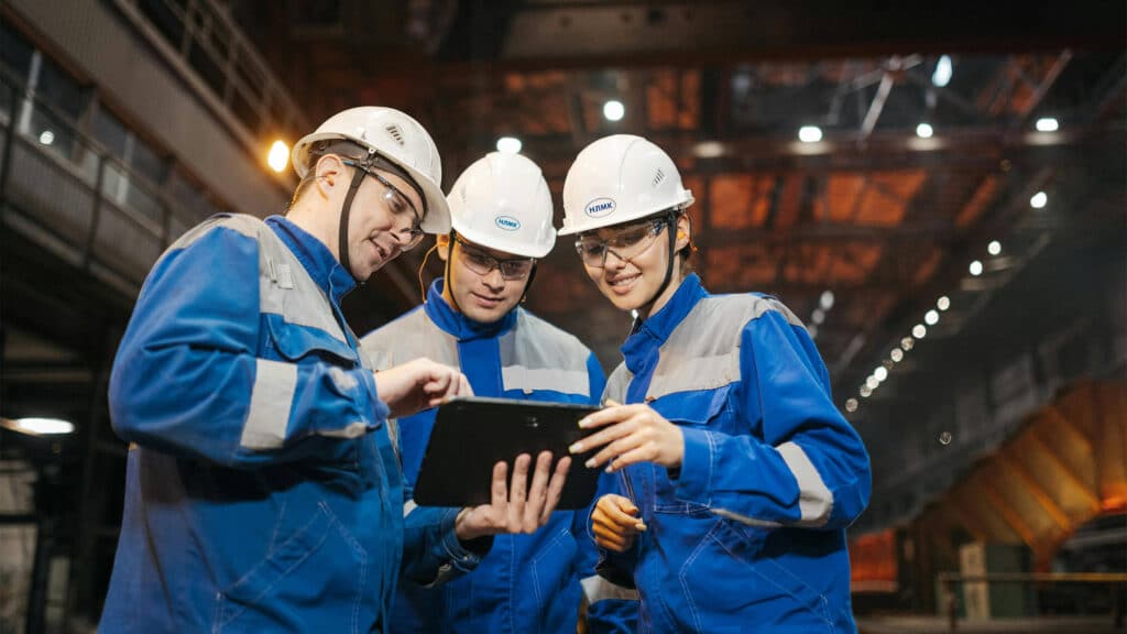 A trio of workers in safety equipment huddle around a table in a warehouse facility.