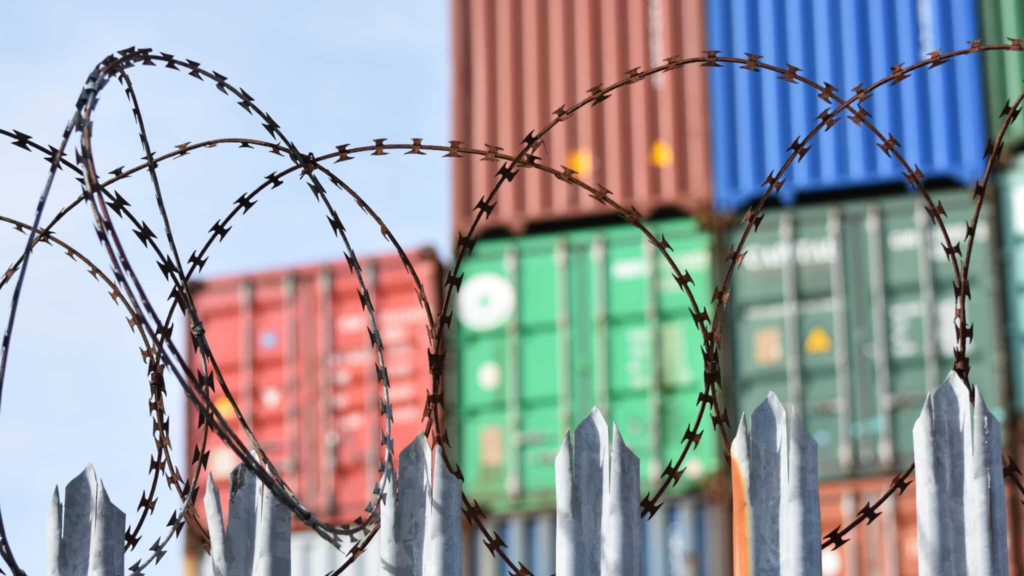 Barbed Wire Fence With Pointed Tops In The Foreground, Stacks Of Colorful Shipping Containers Blurred In The Background. Under A Blue Sky, This Secure Industrial Area Underscores The Impact Of Nor