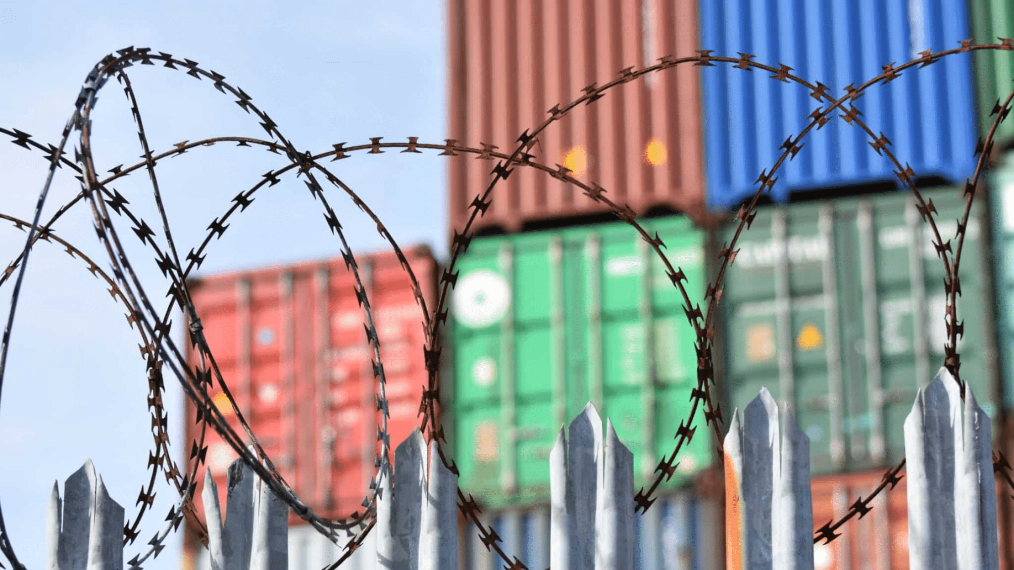 Barbed Wire Fence With Pointed Tops In The Foreground, Stacks Of Colorful Shipping Containers Blurred In The Background. Under A Blue Sky, This Secure Industrial Area Underscores The Impact Of Nor