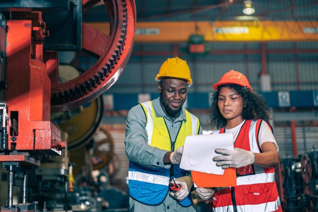 In a factory setting, two industrial workers in protective gear discuss cross training. One, sporting a yellow helmet, points at a document held by the other in an orange helmet and reflective vest, standing near large machinery as they review plans together.