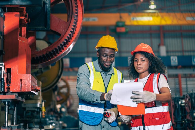 In a factory setting, two industrial workers in protective gear discuss cross training. One, sporting a yellow helmet, points at a document held by the other in an orange helmet and reflective vest, standing near large machinery as they review plans together.