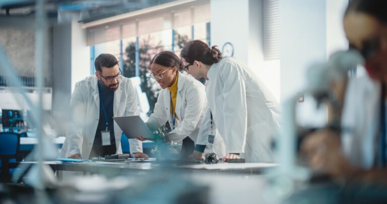 Three scientists in lab coats gather around a table cluttered with equipment, using lab scheduling software on a laptop. The others peer intently at the screen, strategizing their next steps. A bright, modern laboratory serves as the backdrop to their collaborative efforts.