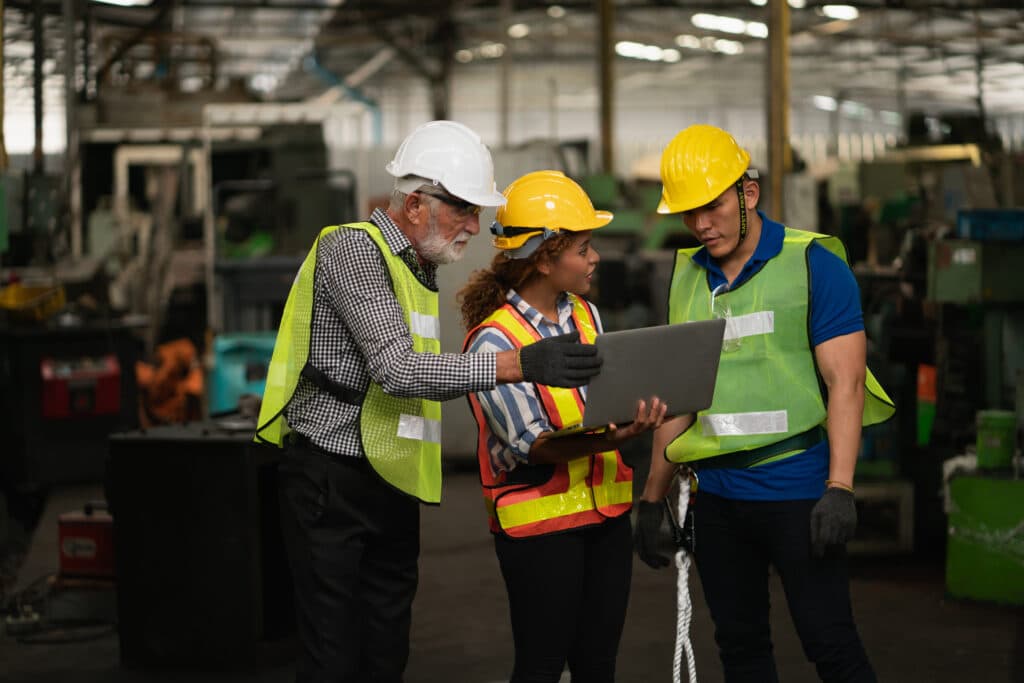 Three engaged factory workers in safety gear and helmets collaborate over a laptop amidst machinery. Their reflective vests and gloves highlight a committed, engaged workforce in this dynamic industrial setting.