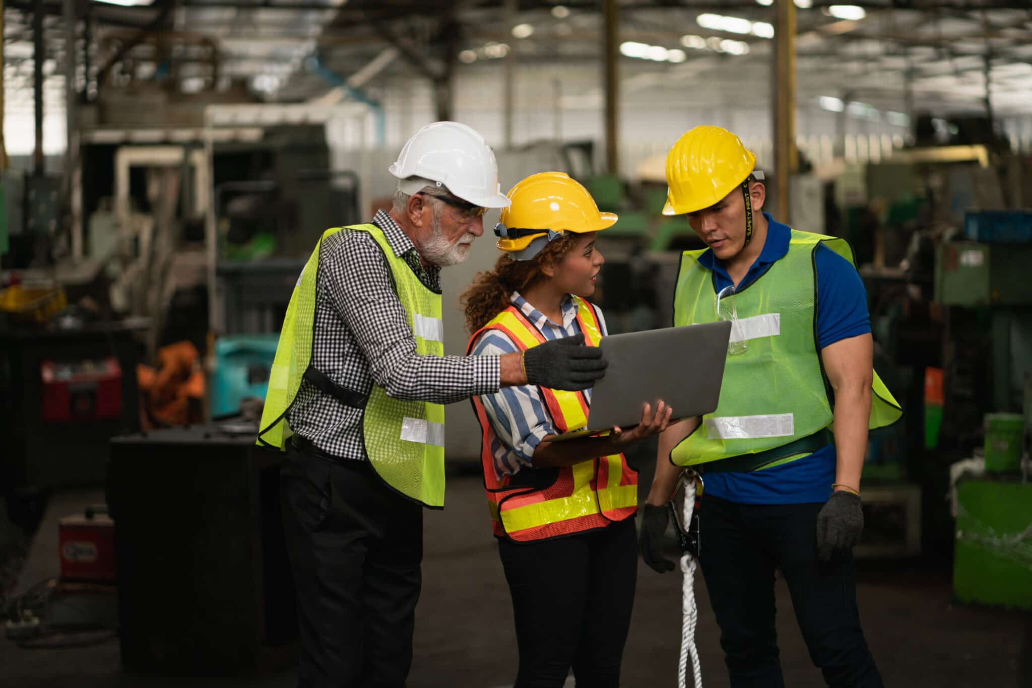 Three engaged factory workers in safety gear and helmets collaborate over a laptop amidst machinery. Their reflective vests and gloves highlight a committed, engaged workforce in this dynamic industrial setting.