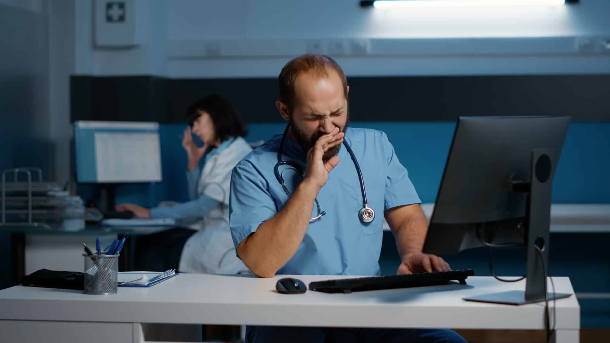 A tired healthcare professional in scrubs yawns at a desk, managing fatigue while working on a computer. In the background, another healthcare worker is on the phone near a glowing screen. The setting is an office or clinic.