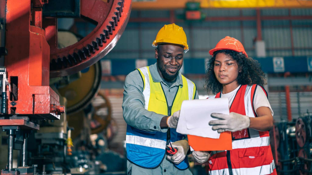 In A Factory Setting, Two Industrial Workers In Protective Gear Discuss Cross Training. One, Sporting A Yellow Helmet, Points At A Document Held By The Other In An Orange Helmet And Reflective Ves