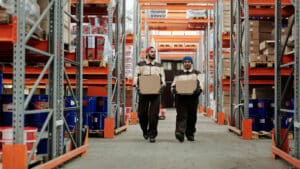 Two factory workers carry boxes through a warehouse.